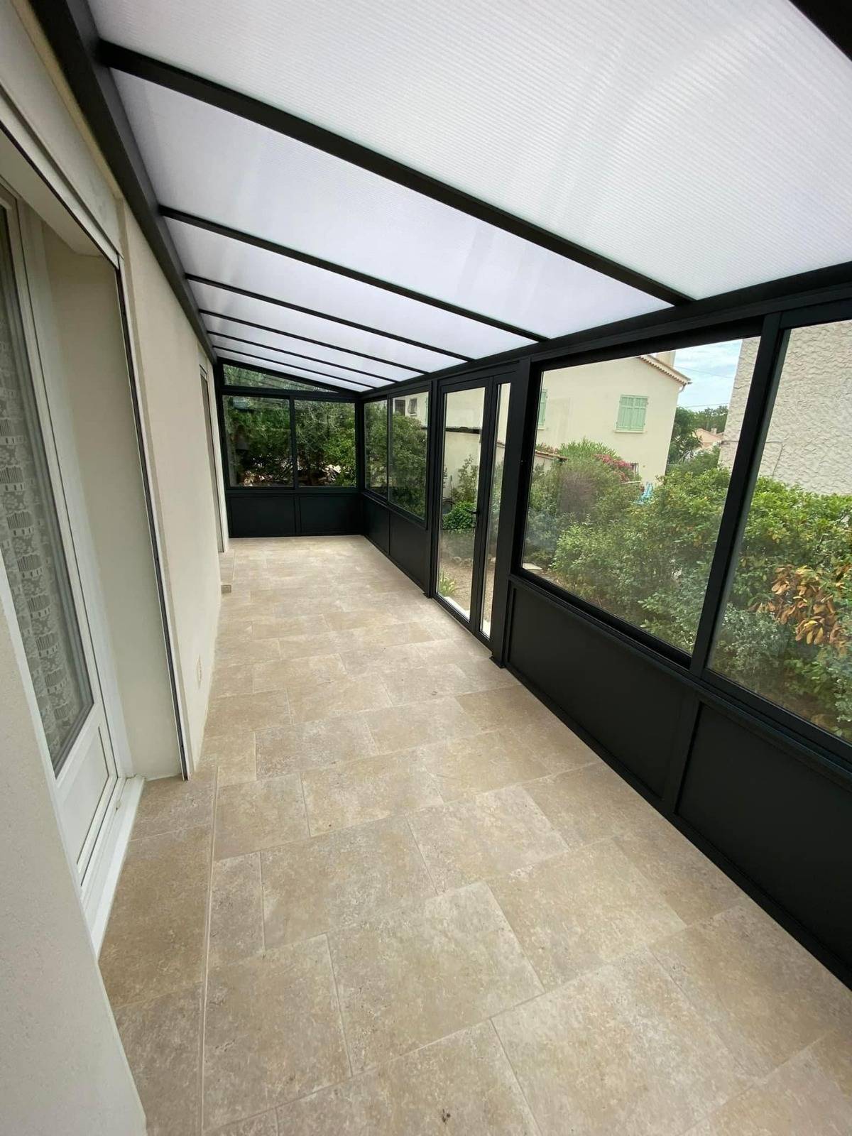 Vue intérieure d’une véranda sur mesure réalisée à Vitrolles par Green House Menuiserie : structure aluminium noire, toiture opaline, sol en pierre naturelle.