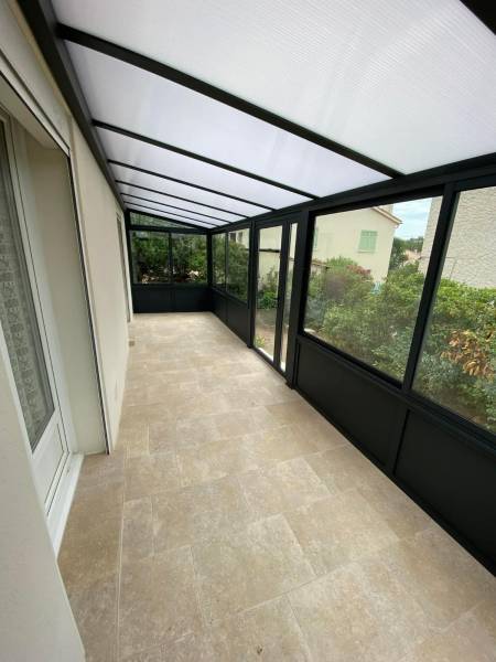 Vue intérieure d’une véranda sur mesure réalisée à Vitrolles par Green House Menuiserie : structure aluminium noire, toiture opaline, sol en pierre naturelle.